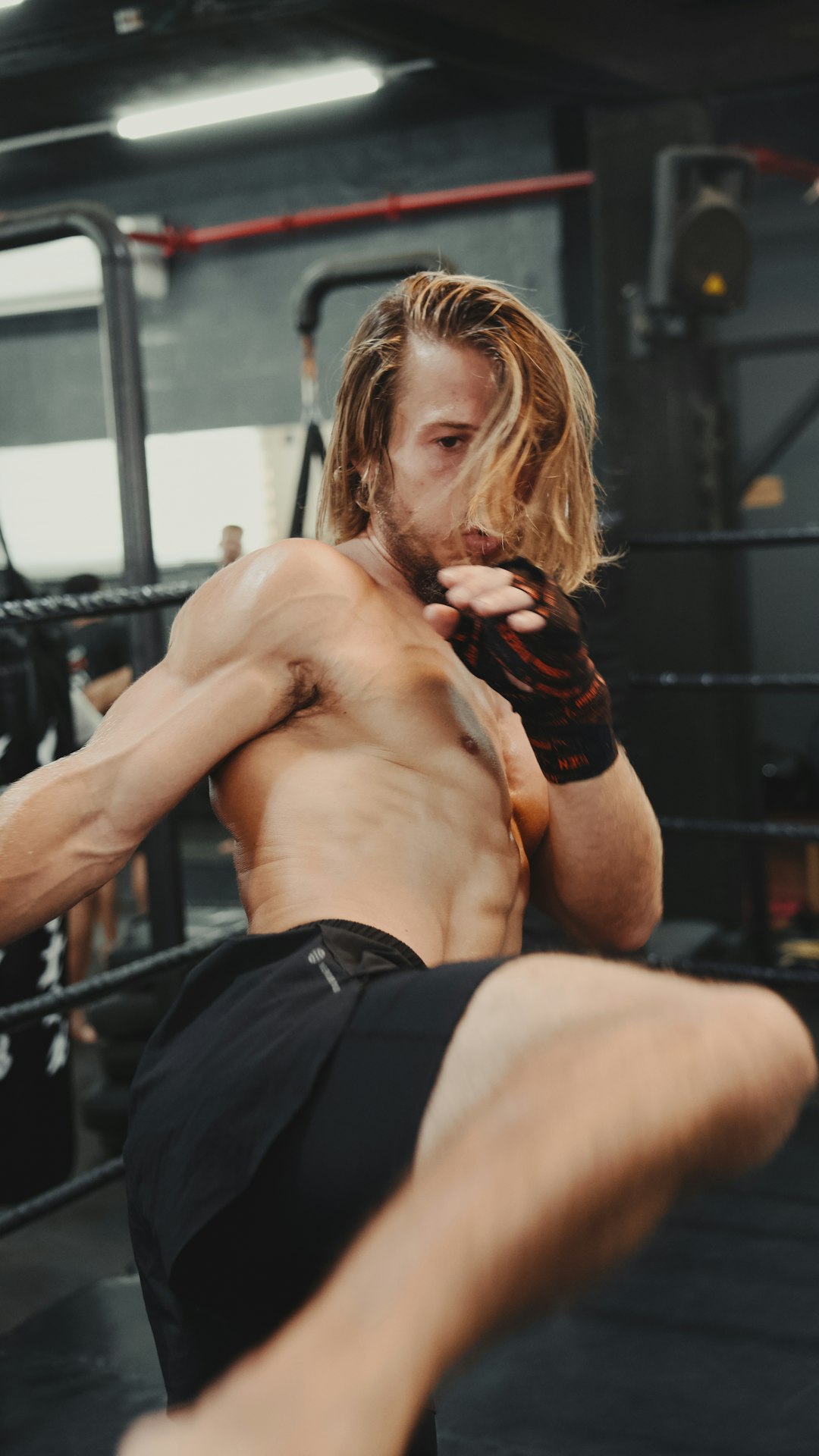 The image shows a person in a gym setting performing a high kick, likely during a martial arts training session. The individual is shirtless, wearing black shorts and gloves, and appears to be in the middle of a dynamic movement, showcasing physical strength and flexibility. The background includes gym equipment and other individuals, indicating an active training environment. This image is interesting and relevant as it captures the intensity and dedication involved in martial arts training, highlighting the physical prowess and discipline required in combat sports.